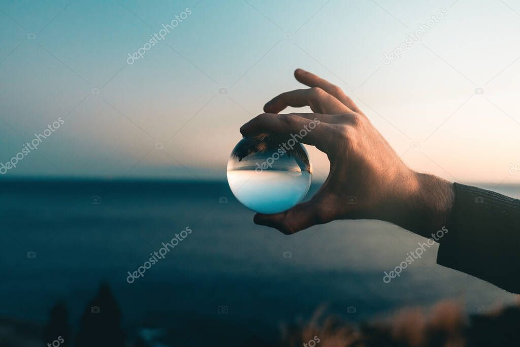 hand holding crystal glass ball during sunrise over sea landscape 