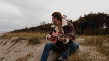 Genç adam sahilde oturuyor ve gitar çalıyor.