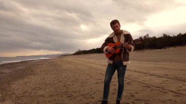 Genç adam sahilde oturuyor ve gitar çalıyor.