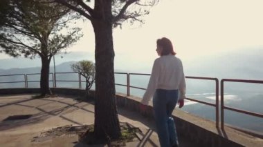 Genç kızıl saçlı kadın gündüz vakti dağlardaki gözlem güvertesinde yürüyor. 