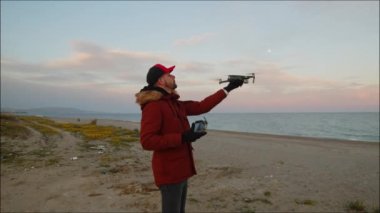 Kışlık elbiseli genç adam güneş doğarken kumsalda dron kullanıyor.
