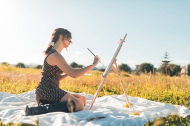 Gün batımında çiçek tarlasında tuvale resim yapan genç bir kadının portresi. 
