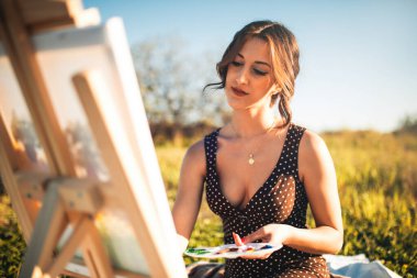 Gün batımında çiçek tarlasında tuvale resim yapan genç bir kadının portresi. 