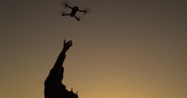 Gün batımında insansız hava aracı uzaktan kumandası kullanan bir adamın silueti. 