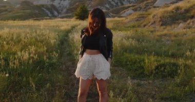 Beyaz şortlu, siyah deri ceketli gün batımında yaz tarlasında yürüyen genç bir kız.