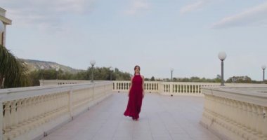 Kırmızı elbiseli genç kız gündüz vakti balkonda yürüyor.