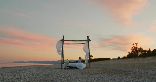 日出时分 身穿蓝色衬衫的小女孩躺在海滩的天篷床上 — 图库视频影像