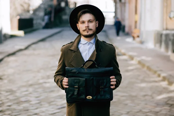 Chipeș tânăr barbă hipster tip în pălărie Fedora pe stradă cu valiză. Retro vintage aspect de moda — Fotografie, imagine de stoc