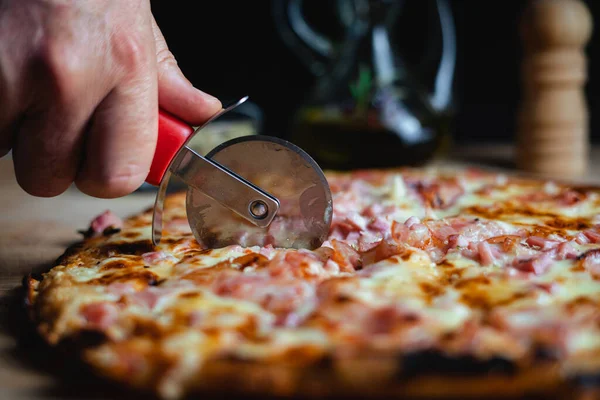 A man cutting a pizza using a pizza cutter. There are oregano, olive oil and pepper in the background. (Horizontal)