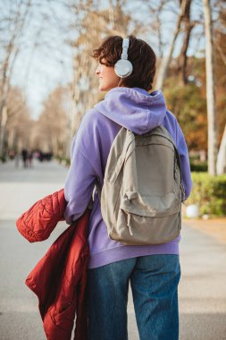 Öğrenci kız müzik dinlerken parkta yürüyor. Beyaz kulaklık takıyor..