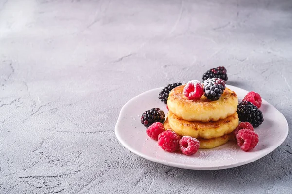 Cottage cheese pancakes and powdered sugar, curd fritters dessert with raspberry and blackberry berries in plate on stone concrete background, angle view copy space