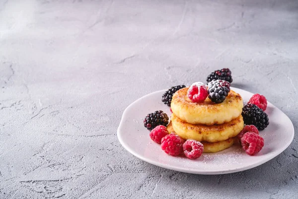 Cottage cheese pancakes and powdered sugar, curd fritters dessert with raspberry and blackberry berries in plate on stone concrete background, angle view copy space