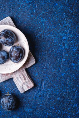 Taze olgun erik meyveleri beyaz ahşap kasede ve mavi soyut arka planda eski kesme tahtası, üst görünüm kopyalama alanı