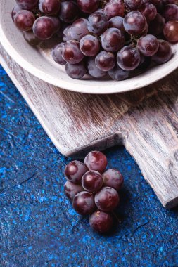 Beyaz ahşap kasede taze üzüm üzümü ve mavi soyut arka planda eski kesim tahtası, açı açılı makro.