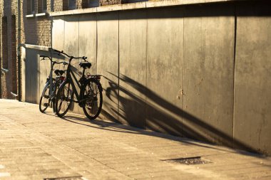 Sıcak akşam ışığında uzun ve dramatik gölgeleri olan bir duvarın yanına park edilmiş iki bisiklet. Şiirsel atmosferli minimalist şehir kompozisyonu.
