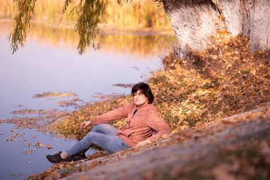 Sonbahar yapraklarının arasında nehir kenarında oturan kadın, rahat kot pantolon ve açık ceket giyiyor, güneşli havanın ve suyun huzurlu yansımasının tadını çıkarıyor..