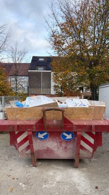 Plastik ambalaj, yalıtım malzemesi ve ahşap panellerle dolu kırmızı bir inşaat çöplüğü konut binalarının yakınındaki çakıl yüzeyinde duruyor..