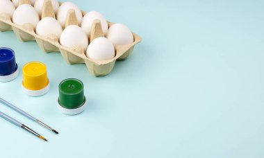 Beyaz yumurtalar, çok renkli boya kutuları ve mavi arka planda boya fırçaları. Paskalya için hazırlanıyorum. Seçici odaklanma. Boşluğu kopyala