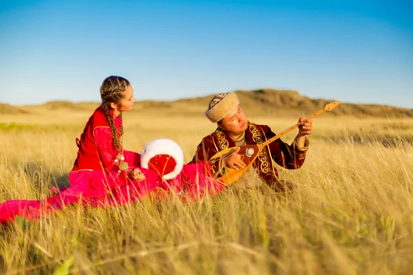 Казахский мужчина и женщина в национальных костюмах в степи играют в домбыру и танцуют — стоковое фото