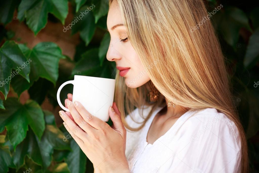 Outdoor portrait of beautiful young blonde holding white cup of tea or coffee. There is place for your logo on the cup.