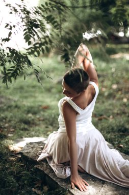 Oturduğu yoga pozu sırasında dengesine ve esnekliğine odaklanan bir kadın yeşil yapraklı sakin bir park ortamında farkındalık ve rahatlama pratiği yapıyor.