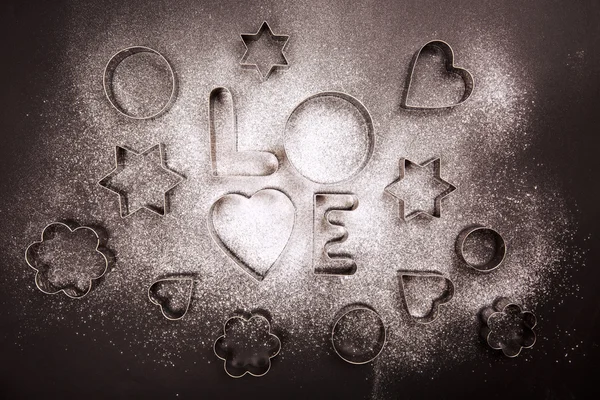 Amor em forma de cortadores de biscoitos e farinha no fundo preto — Fotografia de Stock