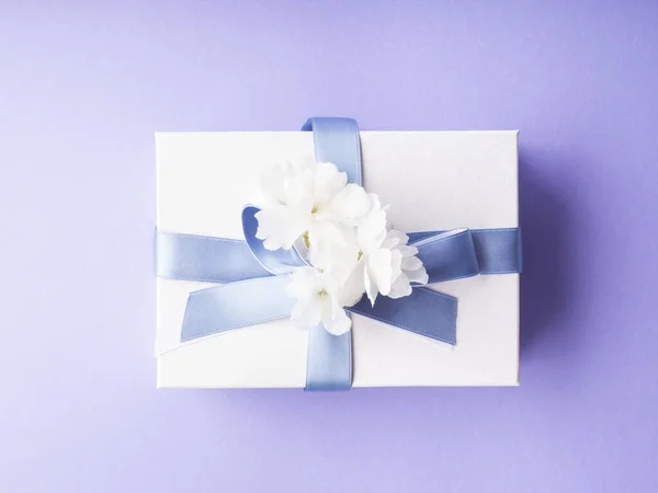 Белая подарочная коробка, обернутая синей лентой и цветами декора на фиолетовом фоне — стоковое фото