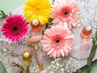 Pembe kuvars yüz tekerlekli gua sha ve serumu mermer masanın üzerinde. Çiçeklerle cilt bakımı kavramı