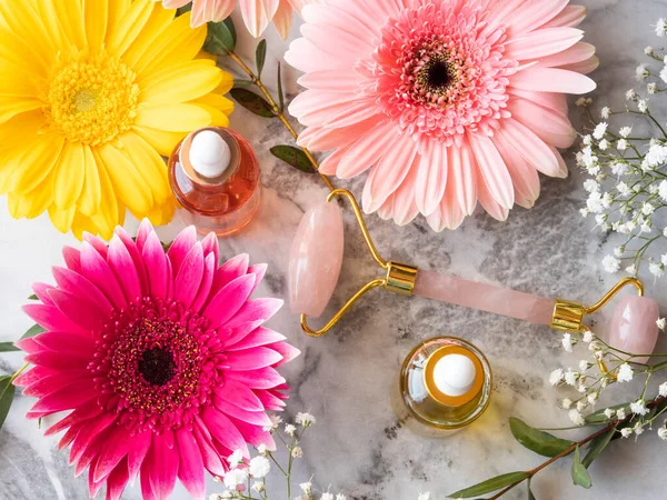 Różowy kwarc twarz rolki gua sha i serum w butelce na marmurowym stole. Koncepcja pielęgnacji skóry z kwiatami — Zdjęcie stockowe