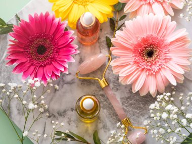 Pembe kuvars yüz tekerlekli gua sha ve serumu mermer masanın üzerinde. Çiçeklerle cilt bakımı kavramı