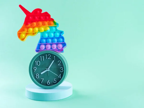 Geometric still life with rainbow unicorn pop it toy, alarm clock and round podium on turquoise
