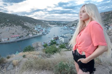 Yazın, bir kız kayaların yanındaki bir dağın tepesinde durur ve arka planda deniz manzaralı bir körfez gezginler için gemileri ve evleri olan bir körfez daha görünür. Burası Kırım 'daki Balaklava kasabası, Sivastopol şehrinden çok uzak değil..