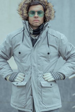 Kış ışığı ceketli bir adam, kafasında bir kapuçon, turkuaz gözlüklü ve elleriyle yan taraflarını tutan kendine güvenen bir bakışla bir duvarın arkasında duruyor.