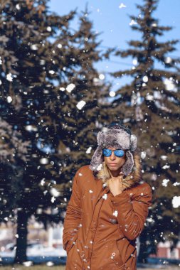 Kış şapkalı ve kürklü bir adam yol boyunca yürür, arka planda yağan karın altında bir köknar ormanı
