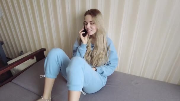 En ung pige taler på en mobiltelefon, sidder på sofaen og ser meget glad ud, har en god samtale – Stock-video