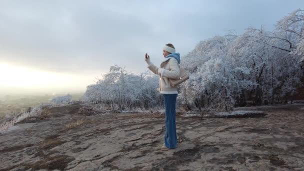 Egy fiatal lány a téli szezonban csinál egy szelfit telefonon egy turista helyen a Mashuk hegyen, a háttérben látható a város és a fák rekedt fagy — Stock videók