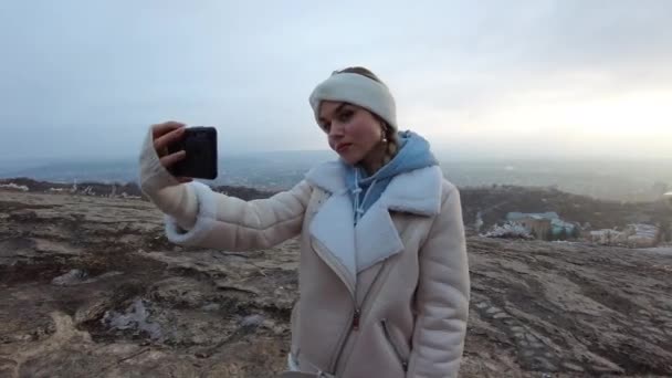 Een jong meisje in het winterseizoen maakt een selfie aan de telefoon in een toeristische plaats op de berg Mashuk, op de achtergrond kunt u de stad en de bomen in de vorst te zien — Stockvideo