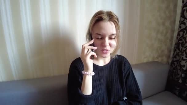 A young girl sits on a sofa in a lotus position and talks on the phone about various household and financial matters with relatives and business partners — Stock Video