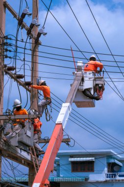 Vinç kamyonlu elektrik ekibi dikey çerçevede mavi gökyüzü arkaplanına karşı elektrik direklerine elektrik sistemleri kurmaya çalışıyor.