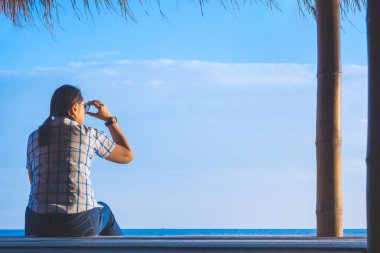 Yazlık evde rahatlayan ve kendinden emin bir jest ve umut dolu bir ifadeyle denize bakan, işaretli gömlekli Asyalı bir kadın..