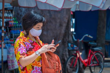 Sahilde yürüyüş alanında akıllı telefon kullanan, koruyucu maskeli, renkli, sıradan bir Asyalı kadın gezgin.
