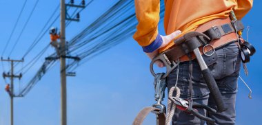 Rear view of electrician with safety belt and work tools is preparing to work on high altitude with blurred background of electrical workers team are working on power poles against blue sky clipart