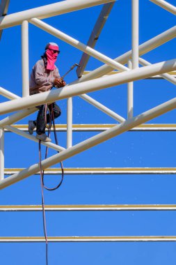 Kaynakçının düşük açılı görüntüsü, inşaat alanındaki depo binasının çatı metal yapısını dikey çerçevede mavi gökyüzüne karşı kaynaklıyor.