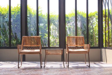 İki rattan sandalye ve büyük cam pencereli küçük bir masa. Güneş ışığı ve yeşil bahçe manzaralı. Ev, kafe veya tatil köyü iç tasarımı için rahat ve rahatlatıcı bir köşe..