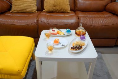 Konforlu oturma odası çay saati renkli badem kurabiyeleri, kek, kruvasan ve beyaz masada kahverengi deri koltuğun ve sarı koltuğun yanında sıcak çay rahatlama ve tatlı aşıkları için sıcak ev atmosferi.