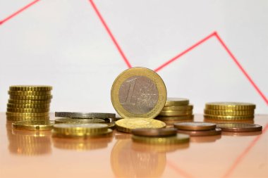 Kapatın. Bir avro ayakta duruyor, artı kontur etrafına saçılmış bozuk paralar ve arka plana istiflenmiş, kırmızı renkli negatif bir grafik.