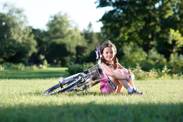 녹색 잔디에 그녀의 자전거 근처에 앉아 어린 소녀. — 스톡 사진