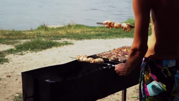 Un uomo prepara il barbecue in riva al lago. Accende la carne del fuoco — Video Stock