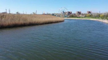 Nehir kıyısı sazlıklarla kaplanmış. Nehir şehir sınırları içinde yer almaktadır. Arka planda bir lunapark var. İnsansız hava aracı ile çekilen video