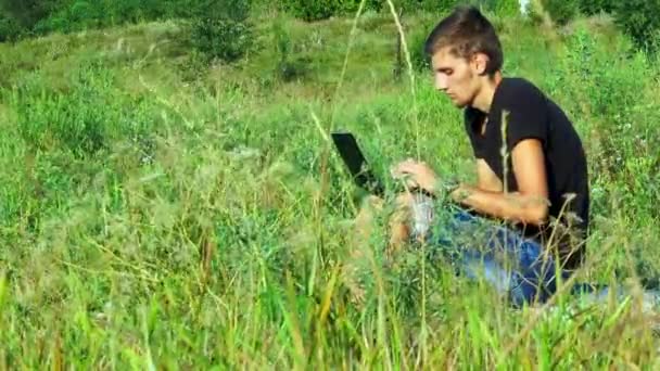 El tipo sentado en la hierba y trabajando en el portátil — Vídeo de stock